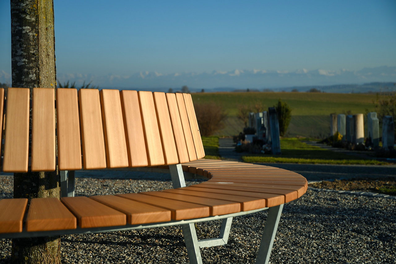 Baumbank rund aus Schweizer Holz, Lärche FSC, Stahl verzinkt im Abendlicht. Hintergrund Alpen und Felder sichtbar